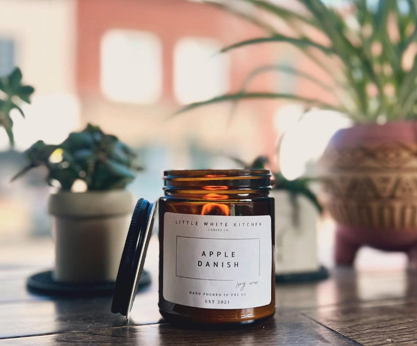 Jar of 'Apple Danish' candle from Little White Kitchen on a wooden surface with blurred plants in the background