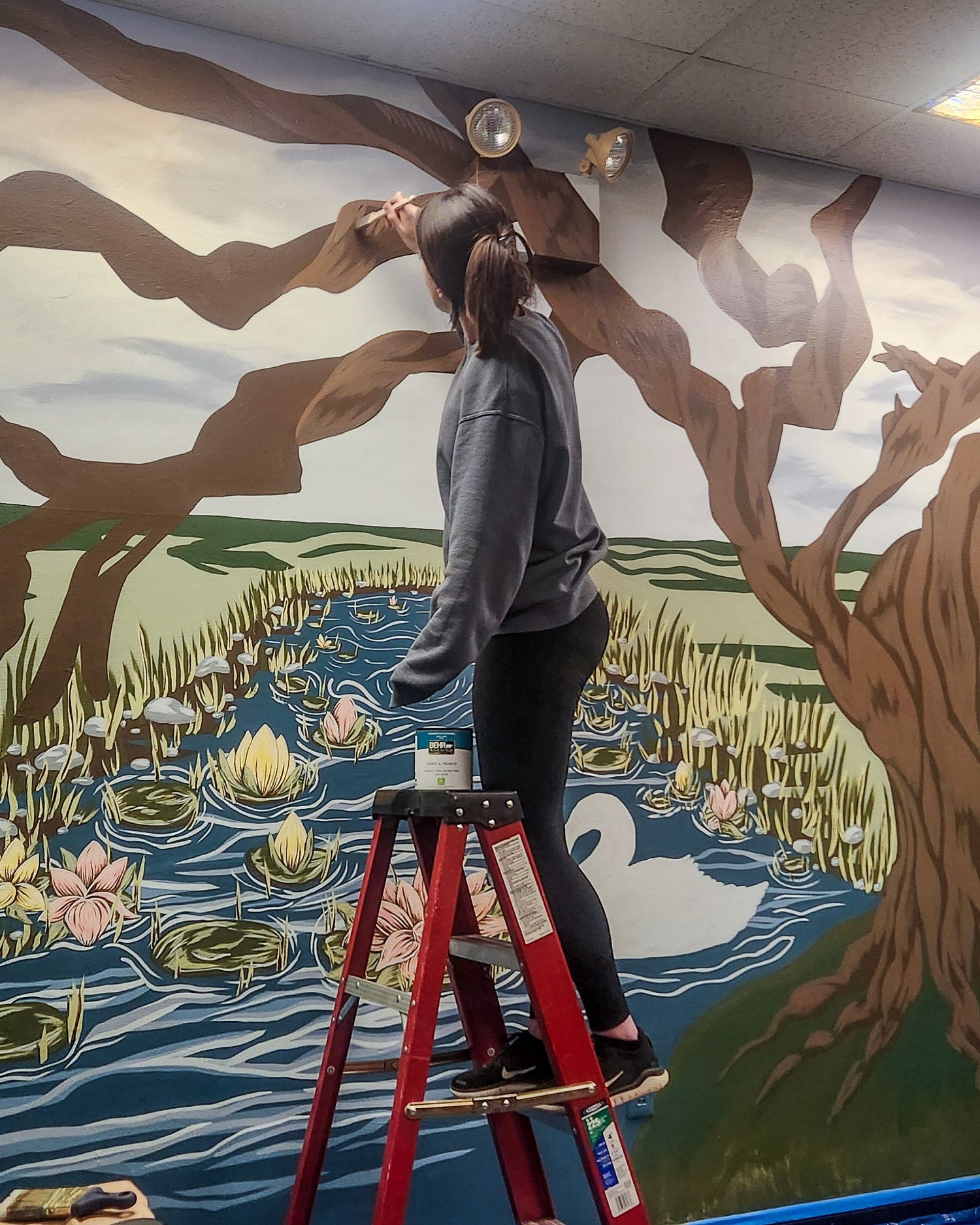 Person painting a large mural of swans and water lilies on a wall.